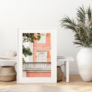 A white matted print sitting on a light colored wood floor, leaning against a white bench and sitting next to palm leaves in a white vase. the photo in the frame depicts a coral pink colored building in Sintra, Portugal, with a white stone balcony and white windows.