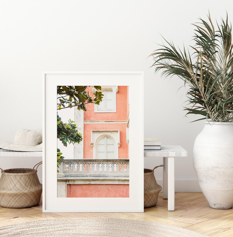 A white matted print sitting on a light colored wood floor, leaning against a white bench and sitting next to palm leaves in a white vase. the photo in the frame depicts a coral pink colored building in Sintra, Portugal, with a white stone balcony and white windows.