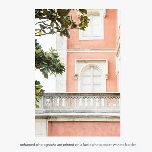 A pink building with white windows and a white balcony. Green leaves from a tree are visible in the upper left corner of the image. Under the photo there is text that reads “unframed photographs are printed on a lustre photo paper with no border.