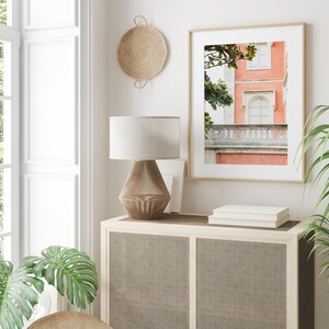 An organic modern living room with a light brown basket hanging on a white wall next to a framed print of a coral-pink building with green trees. A lamp with a white shade and a brown base sits on a wooden cabinet with a woven front.