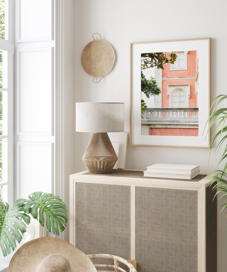An organic modern living room with a light brown basket hanging on a white wall next to a framed print of a coral-pink building with green trees. A lamp with a white shade and a brown base sits on a wooden cabinet with a woven front.