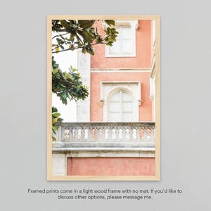 A print of a  coral-pink building with white windows and a carved stone balcony. The building is partially obscured by a leafy tree branch. it is framed in a thin, light colored natural wood frame. the text under the print reads “framed prints come in a light wood frame with no mat. if you’d like to discuss other options, please message me.”
