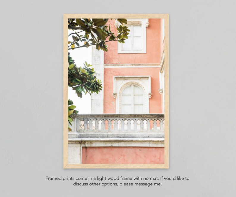 A print of a  coral-pink building with white windows and a carved stone balcony. The building is partially obscured by a leafy tree branch. it is framed in a thin, light colored natural wood frame. the text under the print reads “framed prints come in a light wood frame with no mat. if you’d like to discuss other options, please message me.”