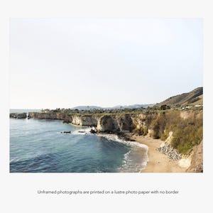 May include: A panoramic view of a rocky coastline with a sandy beach and blue ocean water. The coastline is covered in green vegetation and the sky is a light blue.
