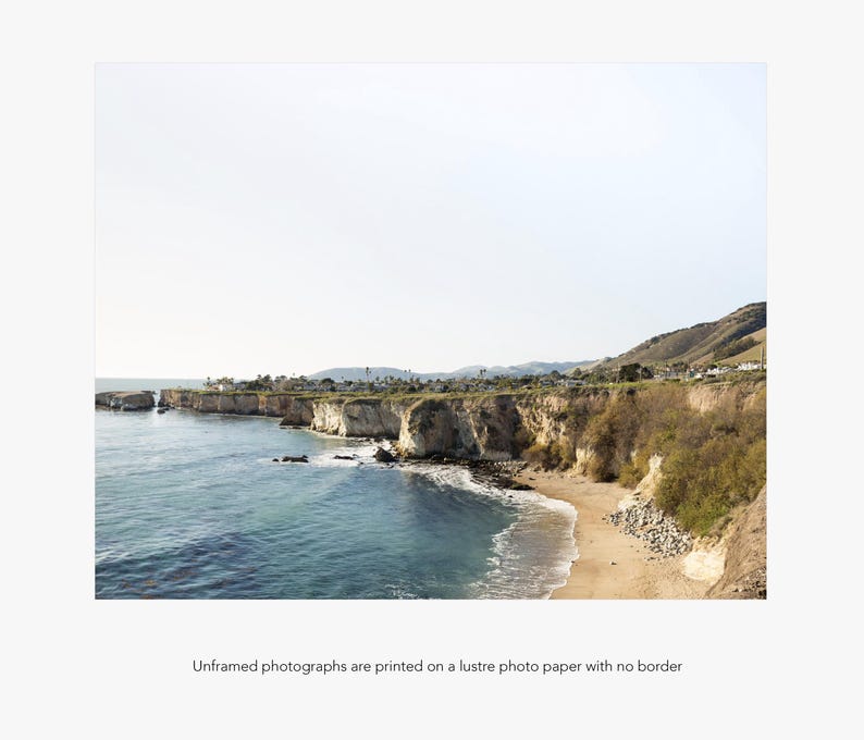 May include: A panoramic view of a rocky coastline with a sandy beach and blue ocean water. The coastline is covered in green vegetation and the sky is a light blue.