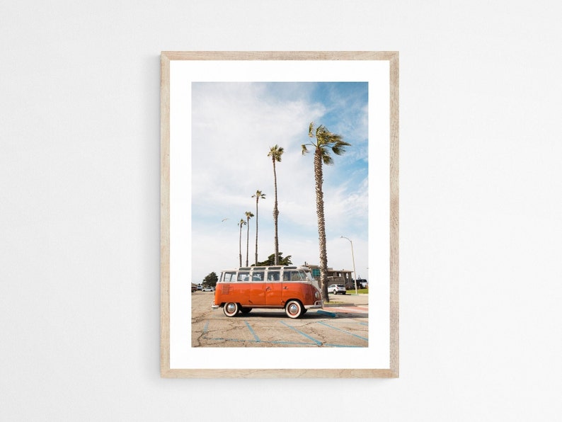 A vertical orientation photograph of an orange vintage Volkswagen bus in a parking lot with palm trees behind it and a seagull flying in the sky.