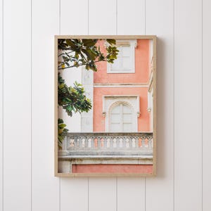 A white shiplap wall with a thin white picture frame hanging on it. In the frame is a photograph depicting a coral colored building with an ornate stone balcony and two windows, with some leaves from a magnolia tree framing the composition.