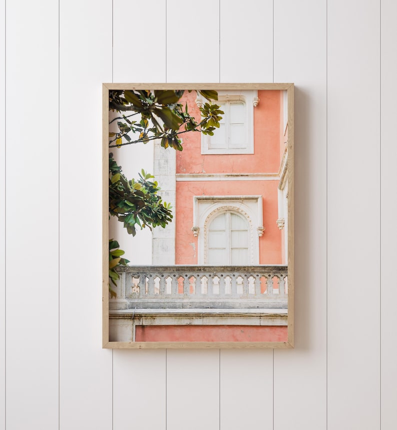 A white shiplap wall with a thin white picture frame hanging on it. In the frame is a photograph depicting a coral colored building with an ornate stone balcony and two windows, with some leaves from a magnolia tree framing the composition.