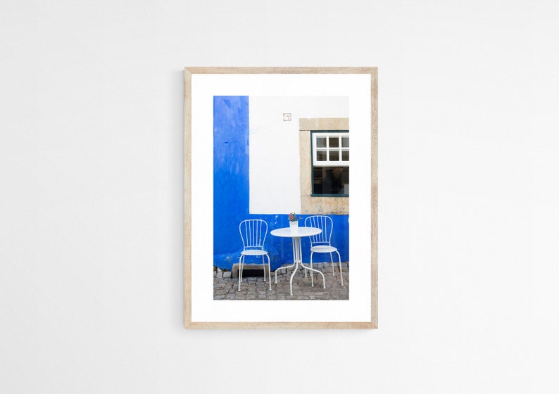 A pair of white iron cafe chairs at a little white table against a bright blue wall with an open window in Obidos, Portugal