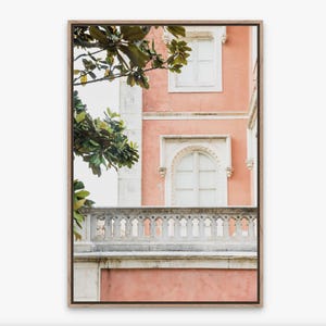A coral- pink building with white windows and a white stone balcony. Green leaves from a tree are visible in the upper left corner of the image. it is printed on canvas and float framed in a thin, light wood frame.