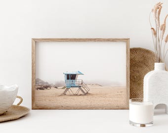 May include: A framed print depicting a light blue lifeguard tower on a sandy beach under a misty sky. The wooden frame surrounds the coastal scene, creating a sense of calm and seaside appeal. Ideal for adding a touch of coastal decor to any home.
