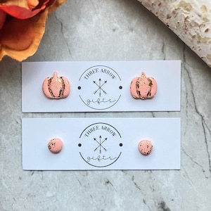 Puede incluir: Dos pares de pendientes de botón en tarjetas blancas con la inscripción Three Arrow Gifts. El par superior muestra calabazas color melocotón con detalles en pan de oro. El par inferior consiste en pequeños pendientes redondos color melocotón.