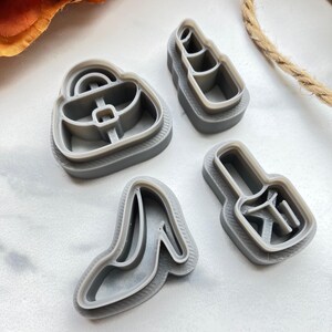 May include: A set of four grey plastic cookie cutters in the shapes of a handbag, a lipstick tube, a high-heeled shoe, and a nail polish bottle. Each cutter features a raised rim and internal structural dividers for creating detailed impressions.
