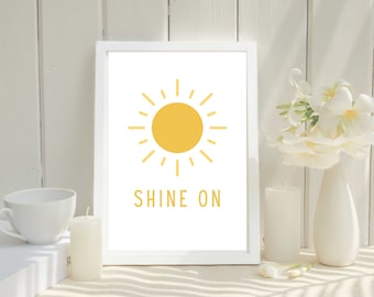May include: A white framed print with a yellow sun and the text "SHINE ON" in yellow. The print is sitting on a white surface with a white candle and a vase of white flowers.