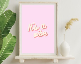 May include: A framed print with a pink background and the text "It's a vibe" in a retro-style font with a yellow outline and a pink shadow.