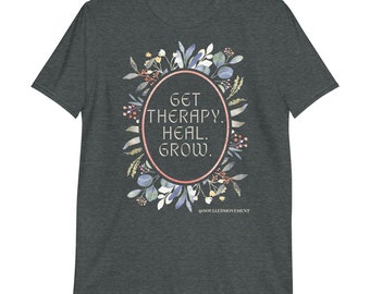 Puede incluir: Camiseta gris oscuro jaspeada con un diseño de corona de flores y el texto "GET THERAPY. HEAL. GROW." en fuente dorada. El texto está dentro de un óvalo rosa. La parte inferior de la camiseta tiene el texto "@SOULLEDMOVEMENT".