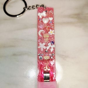May include: Pink glitter nail clipper keychain with decorative embellishments. The rectangular resin piece features white hearts, flowers, bows, and other charms. A silver chain and keyring are attached.