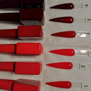 May include: A collection of six nail polish shades, ranging from deep burgundy to vibrant red. Each bottle is paired with a swatch on a clear plastic stick, showcasing the color and finish. The nail polish bottles have a rectangular shape with matching red caps.