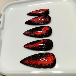 May include: Five red and black cat eye style false nails arranged on a white tray. The nails have a glossy finish with a pointed shape. The red color has a speckled effect.