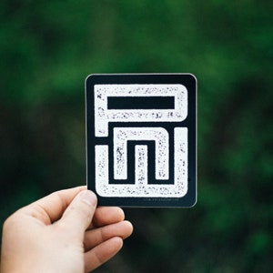 K&ouml;nnte beinhalten: Ein schwarzer Aufkleber mit einer wei&szlig;en Grafik eines stilisierten Buchstabens "R" auf einem strukturierten Hintergrund.