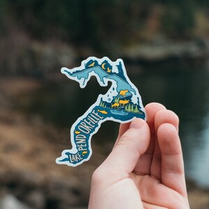 May include: A blue and yellow sticker with a map of Lake Pend Oreille, Idaho. The sticker features a stylized outline of the lake with a few fish, a boat, and a moose. The text "Lake Pend Oreille" is written along the side of the lake.