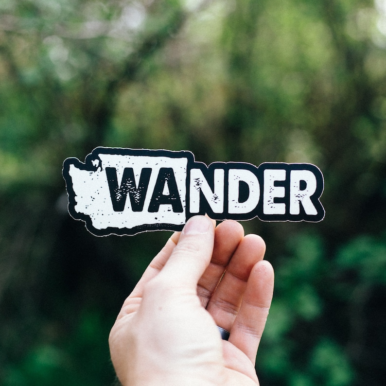 May include: A white and black sticker with the word "WANDER" in bold letters. The word is superimposed on a silhouette of the state of Washington. The sticker is held in a person's hand against a blurred green background.