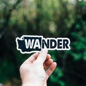 May include: A black and white sticker with the word "WANDER" printed in a distressed font. The sticker is shaped like the state of Washington.