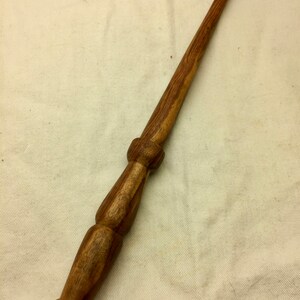 May include: A wooden wand with a brown and dark brown wood grain pattern. The wand has a rounded tip and a thicker handle.
