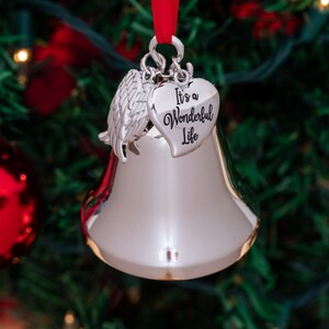 May include: Silver bell Christmas ornament with a heart-shaped charm that reads "It's a Wonderful Life." The ornament has a red ribbon for hanging and is set against a blurred background of a Christmas tree with red ornaments.