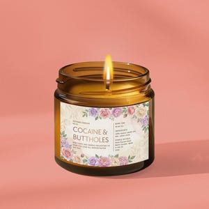 May include: A lit amber glass jar candle with a floral label that reads "Cocaine & Buttholes." The candle has a burning wick and is set against a pink background. The label includes additional text about the candle's ingredients.