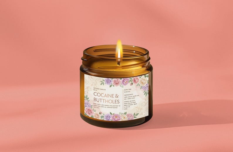 May include: A lit amber glass jar candle with a floral label that reads "Cocaine & Buttholes." The candle has a burning wick and is set against a pink background. The label includes additional text about the candle's ingredients.