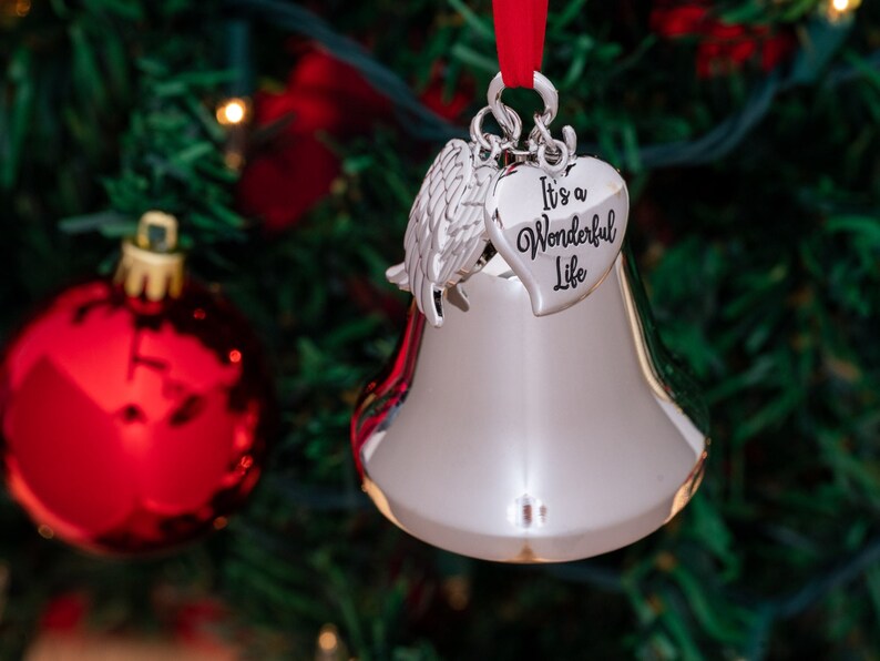 May include: Silver bell Christmas ornament with a heart-shaped charm that reads "It's a Wonderful Life." The ornament has a red ribbon for hanging and is set against a blurred background of a Christmas tree with red ornaments.