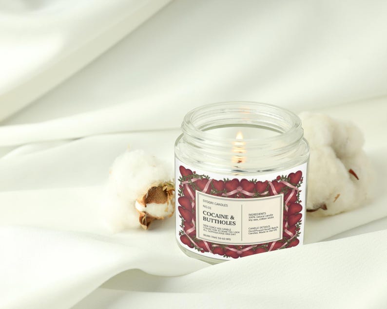 Puede incluir: Una vela encendida en un tarro de vidrio transparente con una etiqueta que dice "Cocaine & Buttholes" rodeada de un dise&ntilde;o de corazones rojos. La vela est&aacute; sobre una superficie blanca junto a capullos de algod&oacute;n.