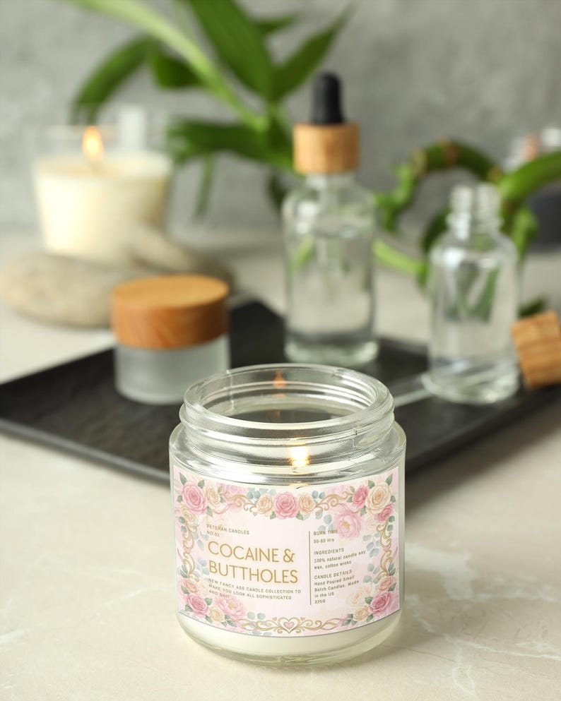 Puede incluir: Una vela encendida en un tarro de cristal con una etiqueta que dice "Cocaine & Buttholes" rodeada de un dise&ntilde;o floral. Otras velas y botellas est&aacute;n en el fondo. La vela forma parte de la colecci&oacute;n "Sleek & Sophisticated".