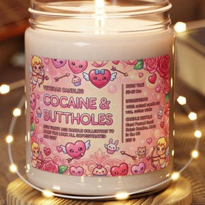 May include: A lit candle in a glass jar with a label that reads "COCAINE & BUTTHOLES." The label features cartoon hearts, cupids, and roses. The candle is on a wooden coaster, surrounded by string lights. The candle weighs 225 g.