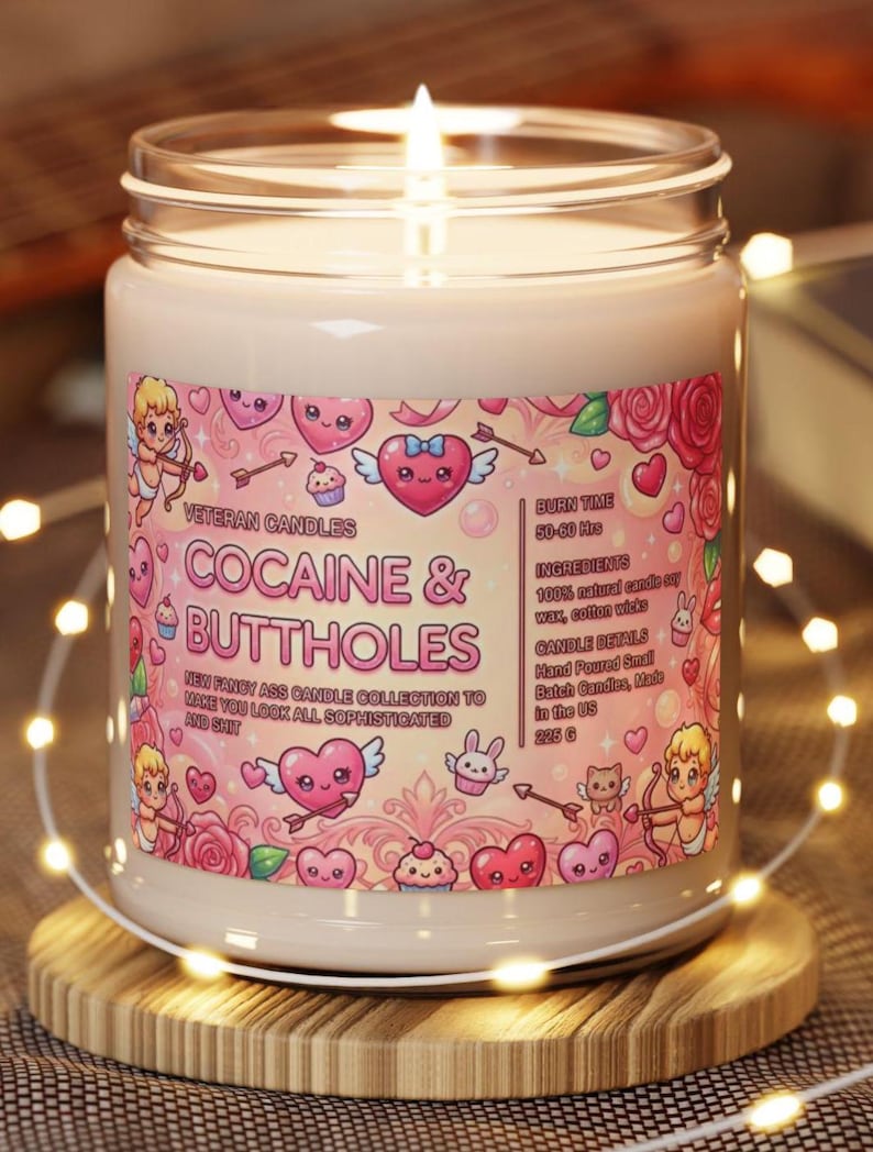 May include: A lit candle in a glass jar with a label that reads "COCAINE & BUTTHOLES." The label features cartoon hearts, cupids, and roses. The candle is on a wooden coaster, surrounded by string lights. The candle weighs 225 g.