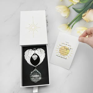May include: A memorial gift set in a white box, featuring a silver angel wing ornament with a crystal prism and a card. The card reads "There are some who bring a light so bright that even after they have gone the light remains."