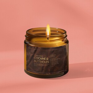 May include: A lit candle in a clear glass jar with a brown label. The label reads "COCAINE & BUTTHOLES" in gold text. The candle is set against a pink background.