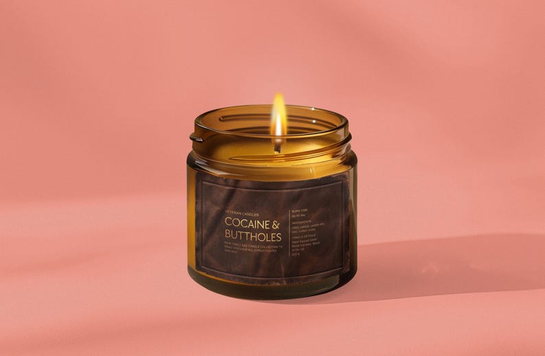 May include: A lit candle in a clear glass jar with a brown label. The label reads "COCAINE & BUTTHOLES" in gold text. The candle is set against a pink background.