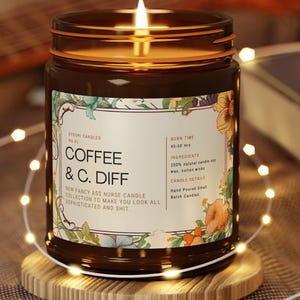 May include: A lit brown glass jar candle with a label that reads "COFFEE & C. DIFF". The label has floral designs and text that says "New Fancy Ass Nurse Candle Collection". The candle sits on a wooden coaster and is surrounded by fairy lights.