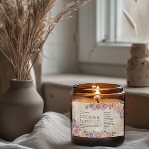 May include: A lit candle in a brown glass jar with a floral label that reads "COCAINE & BUTTHOLES". The candle sits on a white cloth near a window, with a vase of dried plants in the background.
