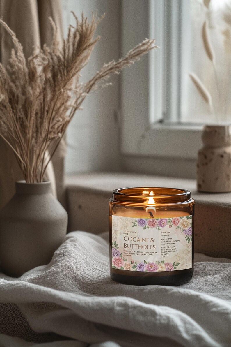 May include: A lit candle in a brown glass jar with a floral label that reads "COCAINE & BUTTHOLES". The candle sits on a white cloth near a window, with a vase of dried plants in the background.