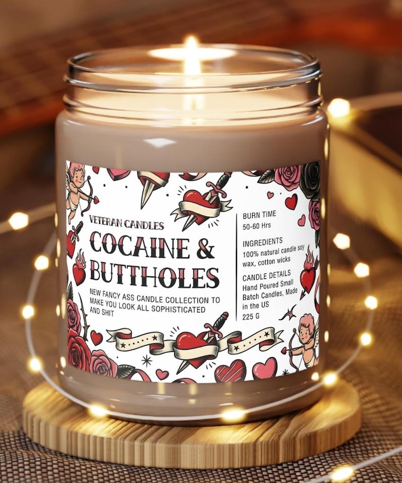 Peut inclure: Une bougie allum&eacute;e dans un bocal en verre avec une &eacute;tiquette indiquant "COCAINE & BUTTHOLES". L'&eacute;tiquette pr&eacute;sente des graphiques de style tatouage de c&oelig;urs, de roses et d'un cupidon. La bougie est sur un dessous de verre en bois et entour&eacute;e de guirlandes lumineuses. La bougie p&egrave;se 225 g.