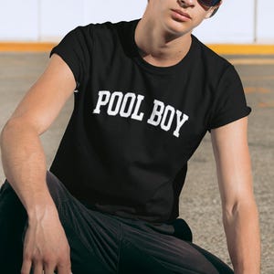 May include: Black t-shirt with the white text "POOL BOY". The shirt is short-sleeved. The person in the image is wearing a grey baseball cap and black sunglasses. The person is also wearing black trousers.