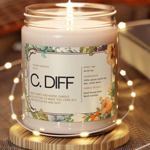 Puede incluir: Una vela encendida en un tarro de cristal transparente con una etiqueta floral. La etiqueta dice "C. DIFF" y "NEW FANCY ASS NURSE CANDLE COLLECTION". La vela está sobre un posavasos de madera, rodeada de luces de cadena. Tiempo de combustión 45-50 horas.