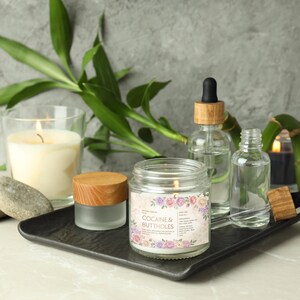 May include: A lit candle in a glass jar with a label that reads "COCAINE & BUTTHOLES" is surrounded by other candles, glass bottles, and bamboo. The items are arranged on a black tray, creating a spa-like aesthetic.
