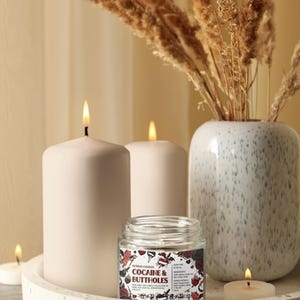 Może przedstawiać: Dekoracyjna aranżacja z zapalonymi świecami, małym szklanym słoiczkiem z napisem "COCAINE & BUTTHOLES" i wazonem z suszonymi roślinami. Świece są białe i cylindryczne, a słoiczek ma kolorową etykietę.