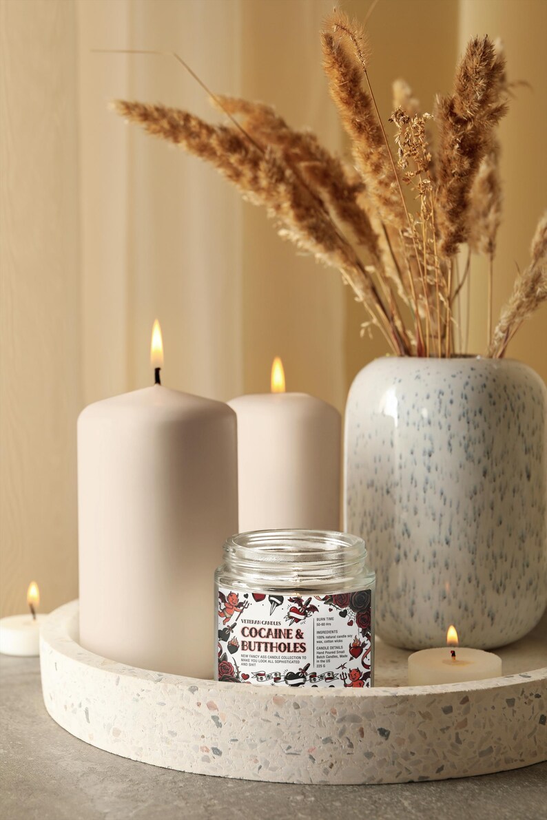 Może przedstawiać: Dekoracyjna aranżacja z zapalonymi świecami, małym szklanym słoiczkiem z napisem "COCAINE & BUTTHOLES" i wazonem z suszonymi roślinami. Świece są białe i cylindryczne, a słoiczek ma kolorową etykietę.