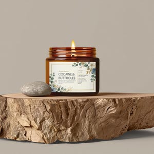 May include: A lit candle in a brown glass jar with a label that reads "COCAINE & BUTTHOLES" is displayed on a wooden slab. The label has floral accents. A small, gray stone sits next to the jar. The candle's flame is bright.