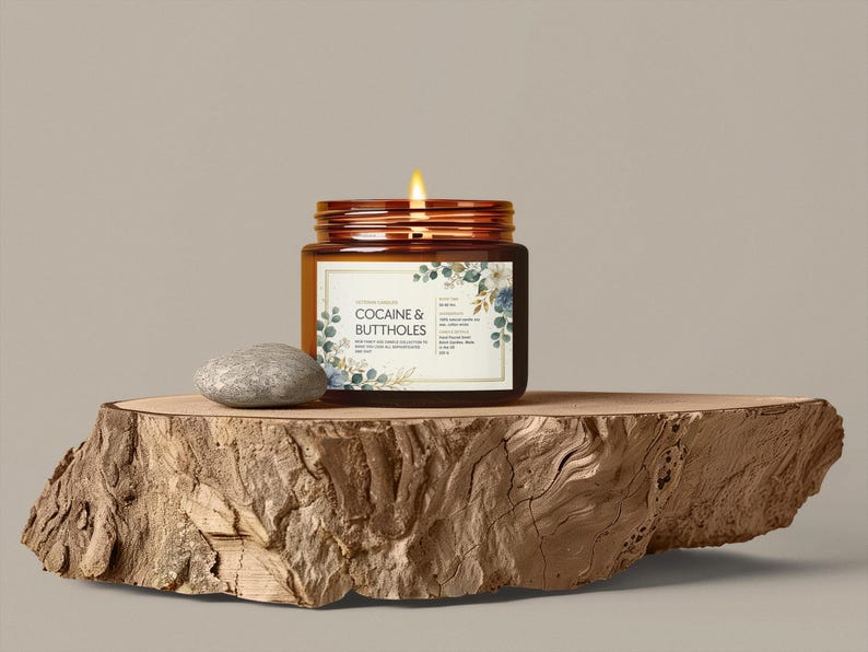 May include: A lit candle in a brown glass jar with a label that reads "COCAINE & BUTTHOLES" is displayed on a wooden slab. The label has floral accents. A small, gray stone sits next to the jar. The candle's flame is bright.